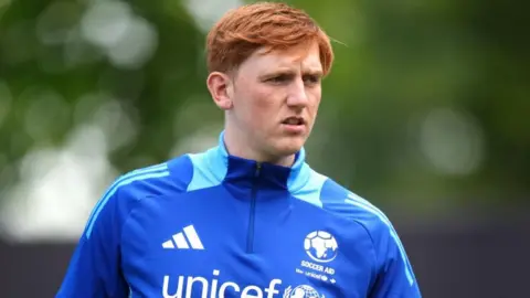 PA Media A man with red hair is wearing a blue Soccer Aid football top with Unicef on the front.