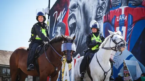 A brown horse and a white one (officially called grey) wearing police visors with mounted police, dressed in protective gear, on their backs. The officers are laughing on a sunny day next to a wall mural of footballer Zaha.