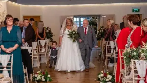 Kimberly, who is wearing a white wedding dress and a veil, is walking down the aisle at her ceremony. She is holding her son Chester's hand and is linking arms with her father. 