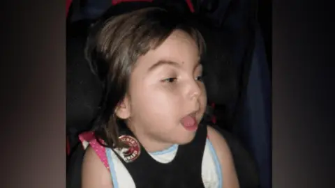 EACH Lucy is sitting in a chair which has a black harness. She has short brown hair and is wearing a pink polka dot dress. 