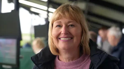 A woman with shoulder length blonde hair, wearing a pink knitted jumper and coat, standing outside smiling at the lens. There are people out of focus behind her.