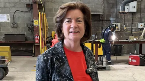 Eluned Morgan is posing towards a camera, wearing a red top under a blue and white jacket.