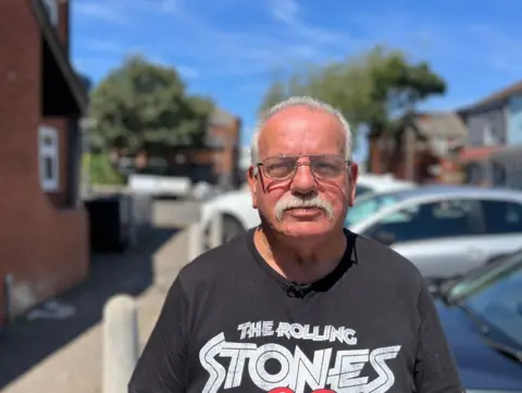 A man with a black T-shirt with the words The Rolling Stones in white is wearing glasses and has a moustache. Properties and cars are in the background.