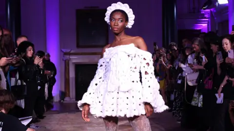 Getty Images A model wearing one of Shonagh's designs at London Fashion Week. The off-the-shoulder mini dress is made from white yarn, has long flowing sleeves and is finished with a large bow on the chest. The model wears a matching crown-style headband. She poses in the purple-lit room in front of a crowd taking photos on their smartphones.