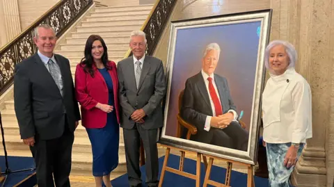BBC Edwin Poots, Emma Little-Pengelly, Peter Robinson and Carol Graham standing by the portrait of Peter Robinson at Stormont. They are standing beside a cream marble staircase. Poots and Robinson are both in suits, while Little-Pengelly is in a blue dress with a red blazer. Carol Graham is standing next to her portrait of a seated Peter Robinson, which is on display on a wooden easel. She is wearing a cream blazer and patterned skirt,