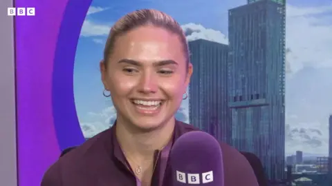 BBC Lauren Knott, who has blonde hair pulled back into a ponytail. speaks into a purple BBC branded microphone