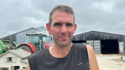 Paul is looking into the camera and smiling slightly. He's wearing a black sleeveless top, and behind him are two huge, corrugated iron cowsheds. In front of the sheds is a red tractor pulling some green machinery. The sky is grey and overcast. 