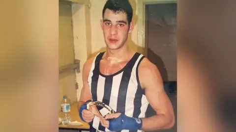 Family photograph Dale Gaucas with black hair in a black and white boxing vest wearing blue boxing wraps holding a boxing trophy after a fight. 