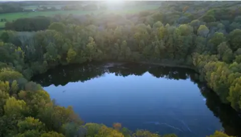 BBC/Wall to Wall A lake is enclosed by green trees, a sheen is visible on the surface