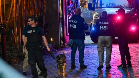 Getty Images At least five agents belonging to Homeland Security Investigations stand on a brick driveway at night in Miami, lit up in blue and red by the lights of an SUV that appears to belong to law enforcement. One of the agents, who is leading a police dog, is wearing dark glasses.