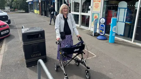 A woman with blonde/grey hair and glasses, standing on a pavement outside a co-operative convenience shop. She has a grey hoodie, black top and purple skirt on. She is holding a black walker with wheels