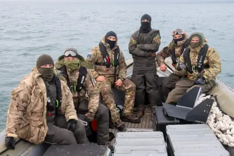Ukrainian Navy Six men dressed in military camouflage and wearing balaclavas to conceal their faces sit in a boat. The expanse of the Black Sea can be seen behind them.