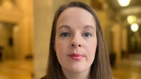 Nuala looking at the camera. She has long straight brown hair and is wearing pink lipstick and a black top with pink flowers on it. 