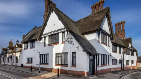 A large white thatched building is on the corner of a street and has a pavement which goes around the front of the building. The windows have red brick ledges, and there are three large chimneys.