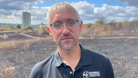 A man with white-grey hair and black glasses. He is wearing a black t-shirt that has a badger logo on the breast and says "Birmingham & Black Country Wildlife Trust". Behind him is a field in a nature reserve completely blackened and charred by fire. There are very few pathway shapes not affected by fire, and they are light brown dead grass. In the distance are high rise buildings