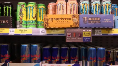 Brightly coloured Monster energy drinks are displayed on a supermarket shelf above rows of Red Bull cans. Several signs show the drinks are on offer as part of a meal deal