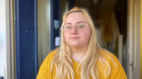 Woman with blonde hair and a yellow jumper stood in doorway