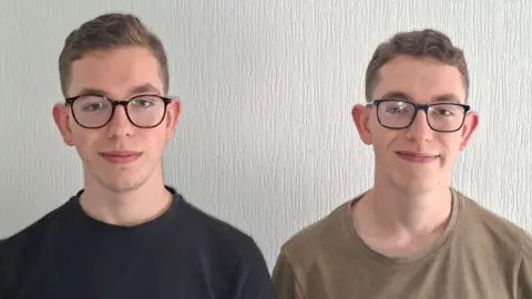 Adrian and Łukasz Kolman Identical twins, Adrian and Łukasz, with short brown hair and glasses standing next to each other smiling. Adrian, on the left, is wearing a black T-shirt while Łukasz wears a light brown top. It is a head and shoulders shot of them both. 