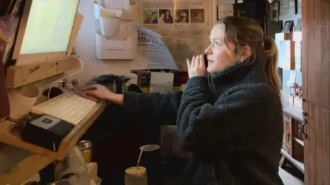 BBC/Seb Cheer A young woman, Isobel, is on the phone while looking at a computer screen with details of table reservations on it.