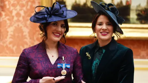 Twin sisters look at the camera. Both are wearing hats. One has a purple dress and has a medal with blue white and red ribbons pinned to it. The other woman wears a dark green jacket over a blouse. She has a crocodile shaped brooch on her jacket.