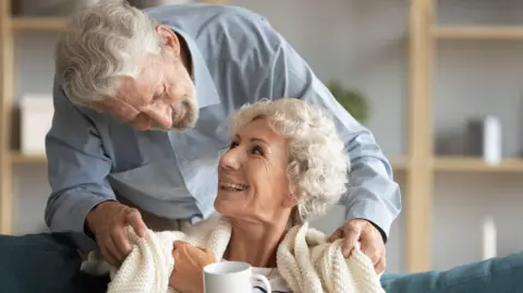 A man with short grey hair looking over the sofa at a woman with short grey curly hair. He is wearing a blue shirt and she has a cream blanket around her as she holds a white mug.