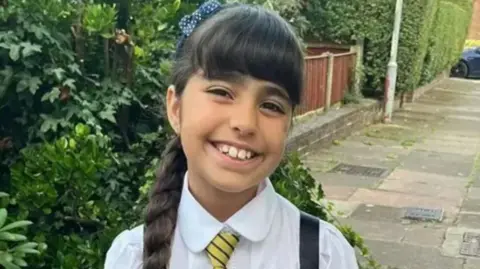 Family Handout Alice Aguiar, wearing her school uniform of a white shirt and a yellow-and-navy striped tie, smiles for the camera. She has long, dark brown hair tied into a plaited ponytail.