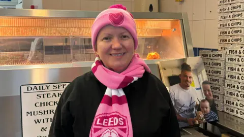BBC/Jack Conlon A lady is standing in a fish and chip shop. She is smiling and wearing a pink Leeds United hat and a pink Leeds United scarf.