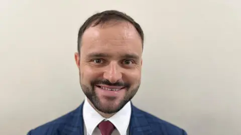 A man with short dark hair and a trimmed beard wearing a white shirt, blue and black suit jacket and dark burgundy tie