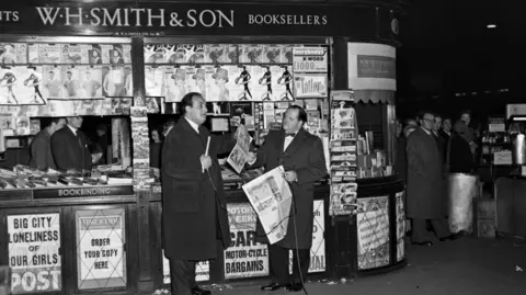 Getty Images Richard Dimbleby filming for French television at Victoria Station, March 1956: a black-and-white image of two men in dark suits standing in front a WH SMITH & SON stall. Two men stand behind the stall. Rows of magazine hang from the top of the stall and newspaper bills are fixed to the bottom of the stall.