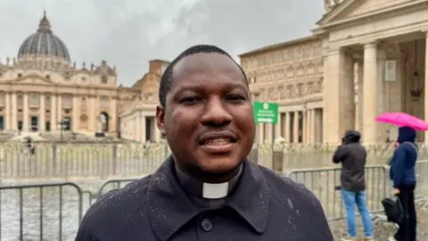 Father Claudio, in a clerical collar, smiles at the camera against the rainy backdrop of Vatican City.