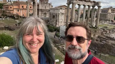 Supplied by Lynda Rousseau A selfie image showing Lynda and Joe Rousseau in front of ancient Roman ruins and columns. Lynda is smiling and has grey hair with bangs and teal streaks. Joe is wearing black sunglasses and has a grey and black beard, and short brown hair.