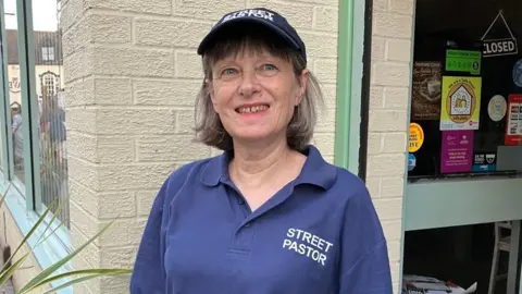 A woman with short grey hair is wearing a blue cap on her head. She is wearing a blue shirt that says Street Pastor. She os standing in front of a white washed brick wall.