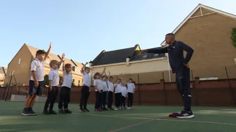 A line-up of primary school children stand on the astroturf pitch in front of Johnny who is coaching. All in white polo t-shirts with their hands up, Johnny is seen pointing to them.