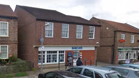 A suburban village street with parked cars in the foreground facing two red brick buildings: Barclays Bank and a Post Office