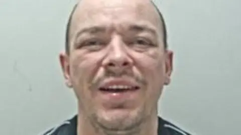 Lancashire Police Police custody image of Jamie Cooper who has short cropped hair, stubble and is smiling at the camera.