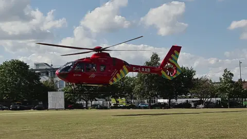A red helicopter with yellow stripes on the tail hovers above the lawn of a large park, about 2 metres above it. You can see an ambulance on the road next to the park and cars parked alongside the fence. There are trees around the edge of the park.