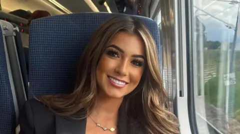 BBC Lily Stroud smiles on a train. She has long, brown hair and dark eye make up.