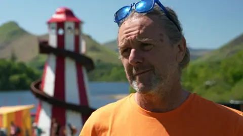 Keswick Mountain Festival organiser Paul McGreal, in an orange t-shirt, looking serious. His shoulder-long grey hair is tied back and there is a pair of blue sunglasses at the top of his head. A red and blue lighthouse-shaped structure with a slide is visible in the background with Derwentwater and Lake District peak further away.