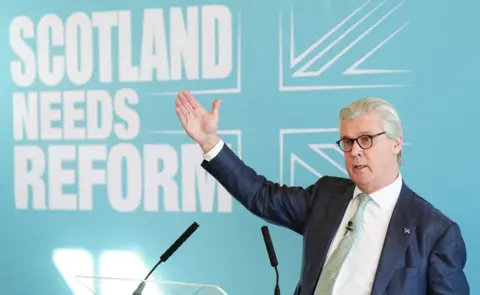 PA Media A grey haired man wearing spectacles and a dark suit, at a news conference with his hand in the air and a Scotland Needs Reform banner in the background