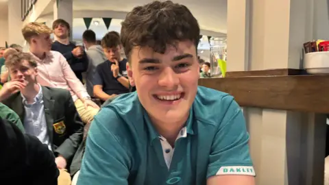 Hamish Johnston has curly dark hair and is smiling at the camera in the bar of Holywood Golf Club. He is wearing a green polo shirt. Behind him other people can be seen watching the golf on TV.