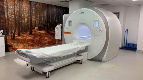 AMANDA WHITE/BBC A picture of a Siemens MRI scanner similar to the one used by the Norfolk and Norwich University Hospital. It has a bed to the front of a donut shaped white tunnel which is used to slide patients into and scan them.