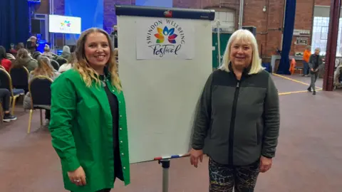 Sian Baxter, an alignment and breathwork coach, stands on the left, wearing a green shirt.  Event organiser Sally Langheim is wearing a dark jacket and the two women are smiling at the camera next to a Swindon Festival A-board. There are people in the background sitting watching a presentation.