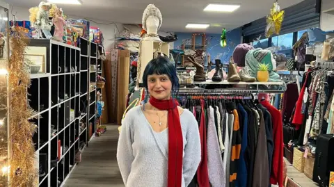 Ashira Zenab with black hair, light blue sweater and dark red scarf in the charity shop with a more contemporary look.