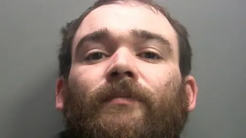 A police mugshot of Blake Stuart. He has brown hair and an unkempt brown beard. He has dark brown eyes and bushy eyebrows. He is staring defiantly at the camera.