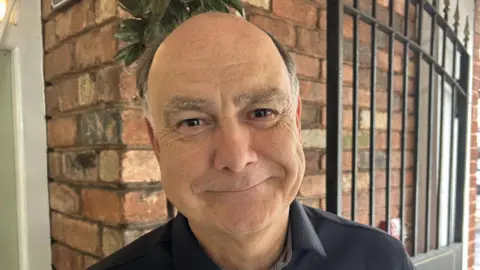 Kevin Allen smiling for the camera. He is a balding man who is clean shaven. There is a brick wall and a black gate behind him.