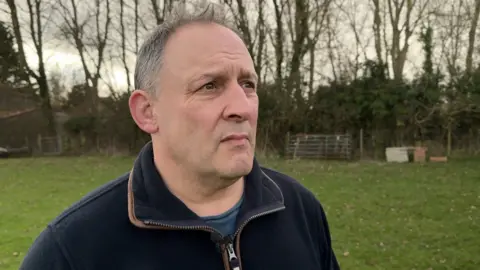 Michael Cook looking over the land he used to own. He is wearing a blue fleece and stood in a green field.