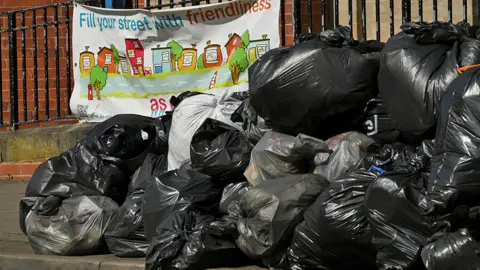 Reuters A pile of black bin bags on the side of the pavement in front of a sign attached to railings reading: "Fill your street with friendliness"