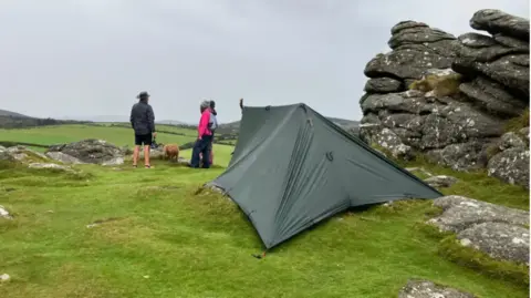 BBC A small tent pitched on uneven grassy ground next to an outcrop of rocks on the flanks of Hound Tor on Dartmoor with three people in the background chatting to each other accompanied by a light brown coloured dog that has its snout pointing downwards as if it is interested in the flora and fauna of the beauty spot.