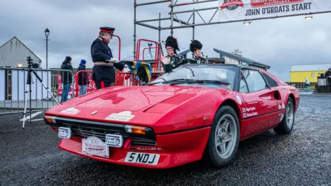 Susie Mackenzie/SDM Photography A sporty red car gets ready to depart while watched by a man in uniform and two pipers. It is raining and the car's bonnet is wet.