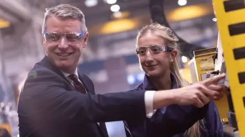 Babcock International Luke Pollard left in a suit and safety goggles pushing a button in a factory setting, he is next to a female engineer who is wearing overalls and safety goggles.